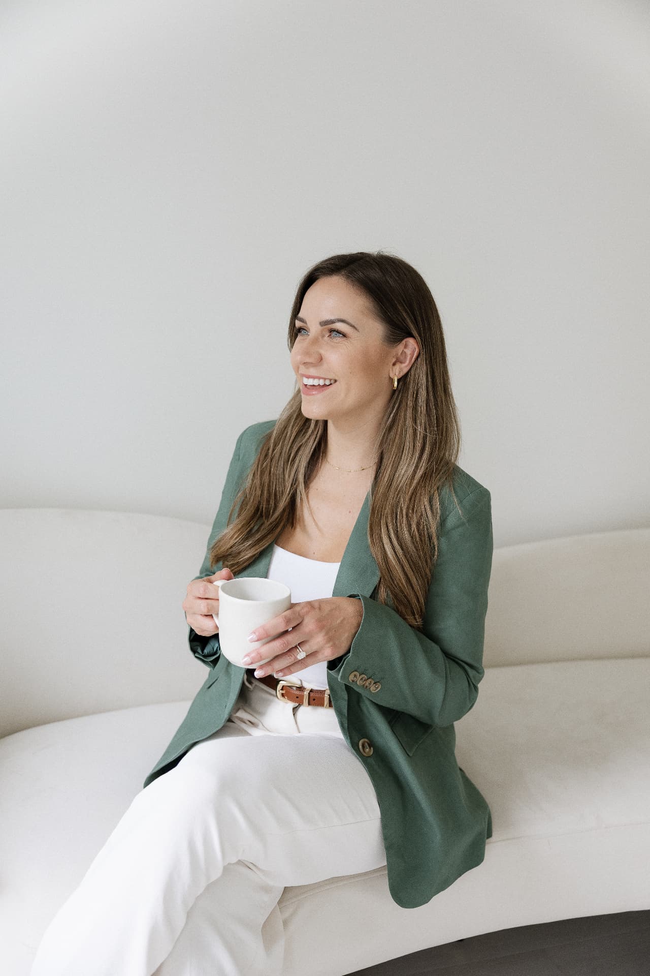 Alyssa seated on a cream sofa in a green blazer holding a mug